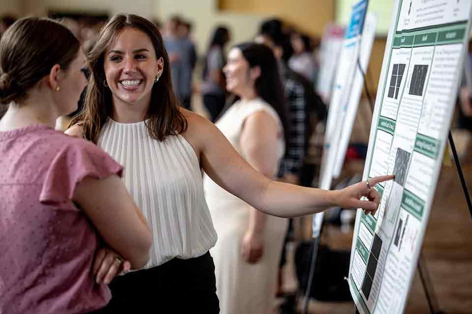 A senior smiles as she shows off her research poster to an attendee of the 2024 Senior Legacy Symposium